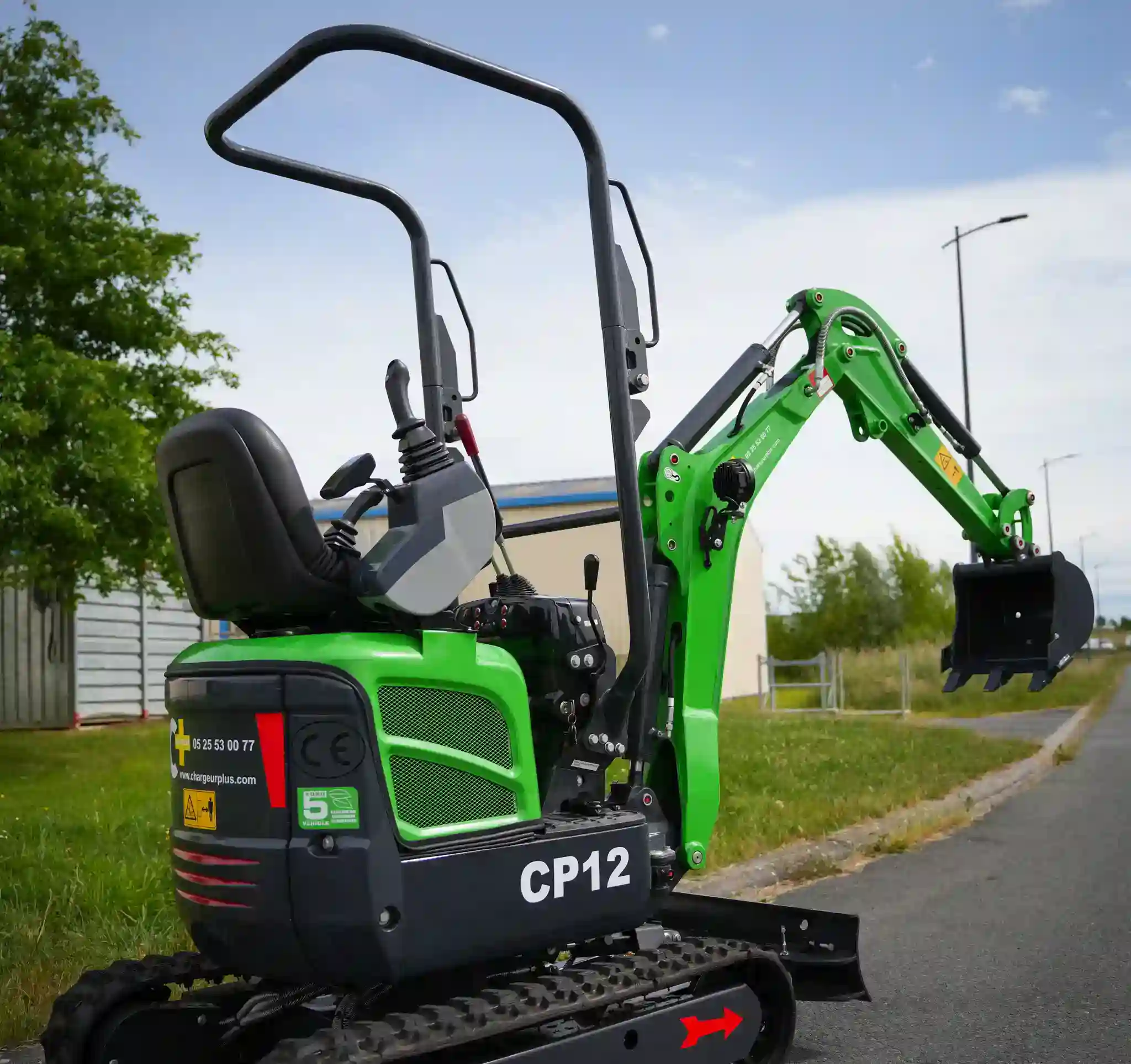 mini-pelle verte CP12 en action sur chantier avec herbe, route et batiment en fond. 