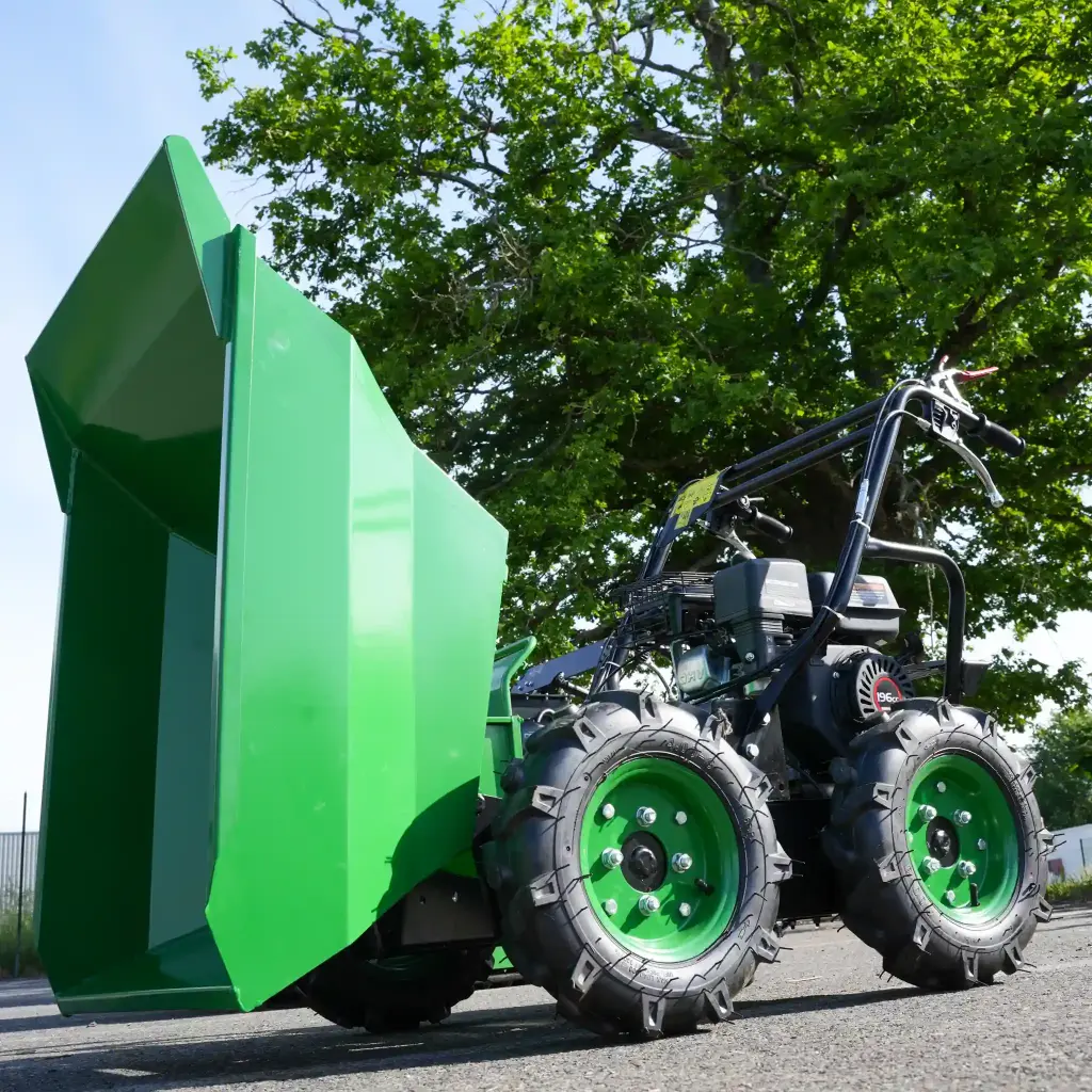 Mini dumper motorisé vert avec benne basculante relevée et roues visibles, stationné sur une route, arbre en arrière-plan