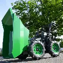 Mini dumper motorisé vert avec benne basculante relevée et roues visibles, stationné sur une route, arbre en arrière-plan