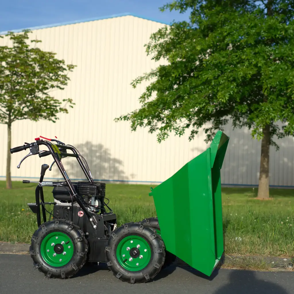 Mini dumper motorisé vert vu de profil, benne en position de déversement et roues visibles, stationné sur route avec bâtiment et arbres en arrière-plan