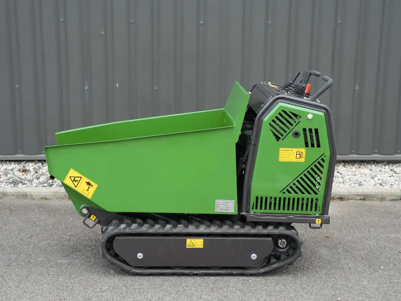 Vue latérale d’un mini dumper vert à chenilles, avec benne basculante, moteur caréné et pictogrammes de sécurité visibles sur fond de mur gris.