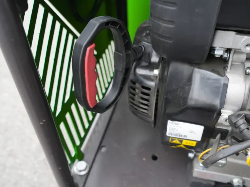 Poignée de lanceur à rappel rouge et noir d’un moteur de dumper, avec détails du carter moteur noir et grille de protection verte en arrière-plan.
