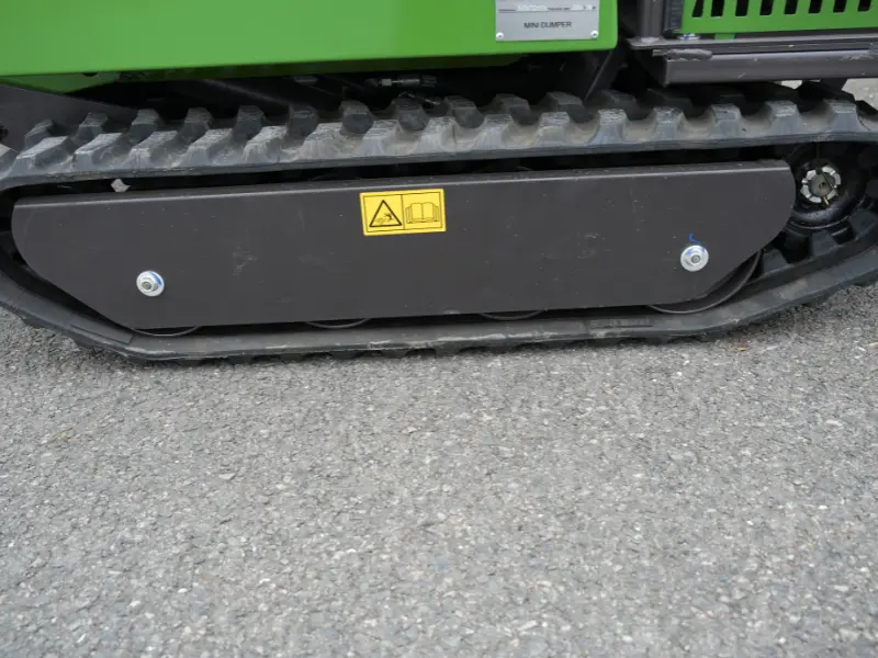 Chenille en caoutchouc d’un dumper vert vue de côté, avec carter de protection noir et pictogrammes de sécurité sur fond bitumé.