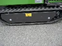 Chenille en caoutchouc d’un dumper vert vue de côté, avec carter de protection noir et pictogrammes de sécurité sur fond bitumé.