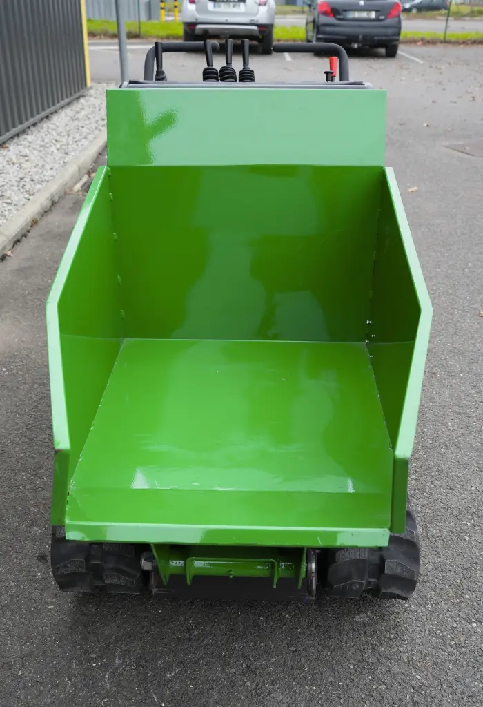 Benne vide d’un dumper à chenilles vert vue de face, avec commandes de pilotage visibles à l’arrière et sol bitumé en arrière-plan.