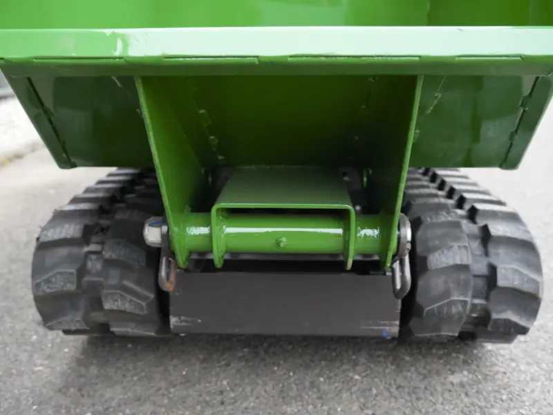 Vue arrière d’un dumper à chenilles vert, centrée sur le châssis et la fixation de la benne, avec les chenilles caoutchouc en contact avec le sol bitumé.