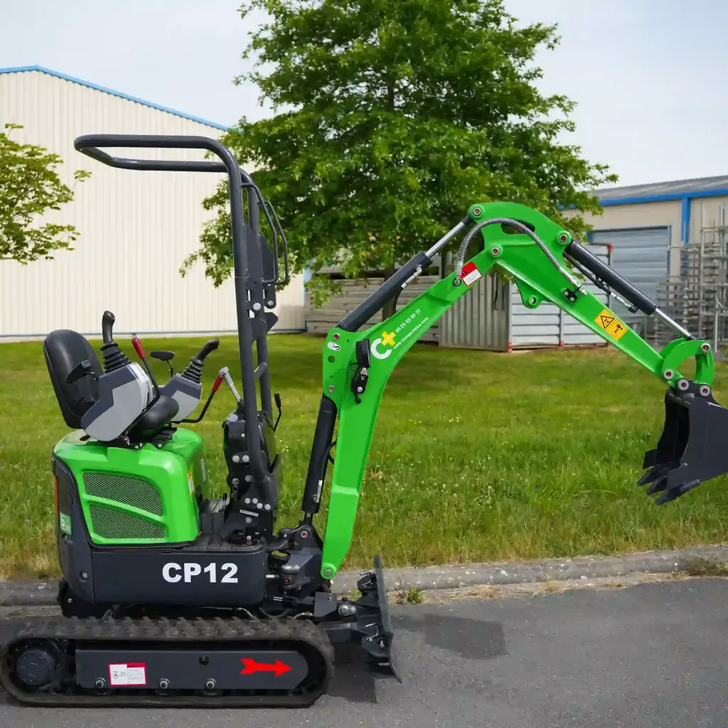 photo de côté d'une mini-pelle CP12 sur un terrain en fond, on apperçoit sur la photo les chenilles de la mini-pelle, les manettes, et un godet au bout du bras de la flèche. Le vérin est inversé