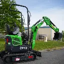 photo de profil d'une mini-pelle CP12 avec un fond en ciel bleu, on apperçoit sur la photo les chenilles de la mini-pelle, les manettes, et un godet ou bout du bras de la flèche. Le vérin est inversé