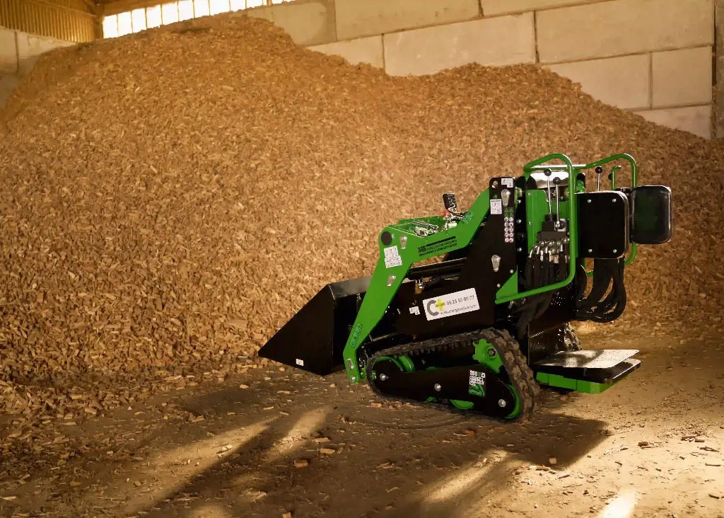 Chargeurplus vous présente le mini chargeur sur chenilles de profil droit, godet abaissé en position de chargement devant un imposant tas de copeaux de bois, démontrant sa maniabilité en espace intérieur agricole.