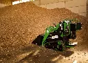 Chargeurplus vous présente le mini chargeur sur chenilles de profil droit, godet abaissé en position de chargement devant un imposant tas de copeaux de bois, démontrant sa maniabilité en espace intérieur agricole.