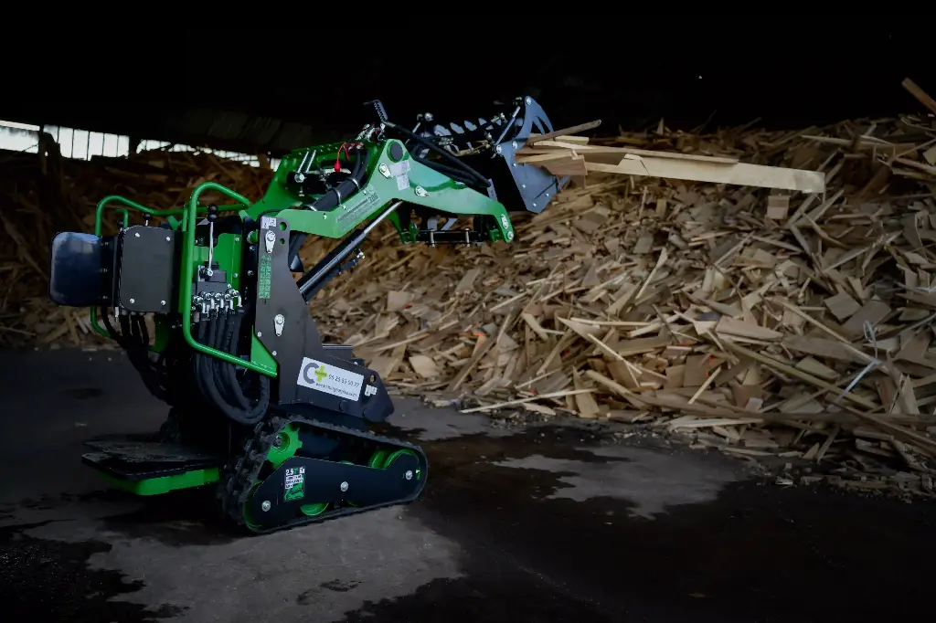 Chargeurplus vous présente le mini chargeur sur chenilles équipé d'un grappin, soulevant des planches de bois devant un important tas de chutes de bois dans un…Chargeurplus vous présente le mini chargeur sur chenilles équipé d'un grappin, soulevant des planches de bois devant un important tas de chutes de bois dans un hangar, démontrant sa polyvalence pour la manutention de matériaux encombrants en espace intérieur.