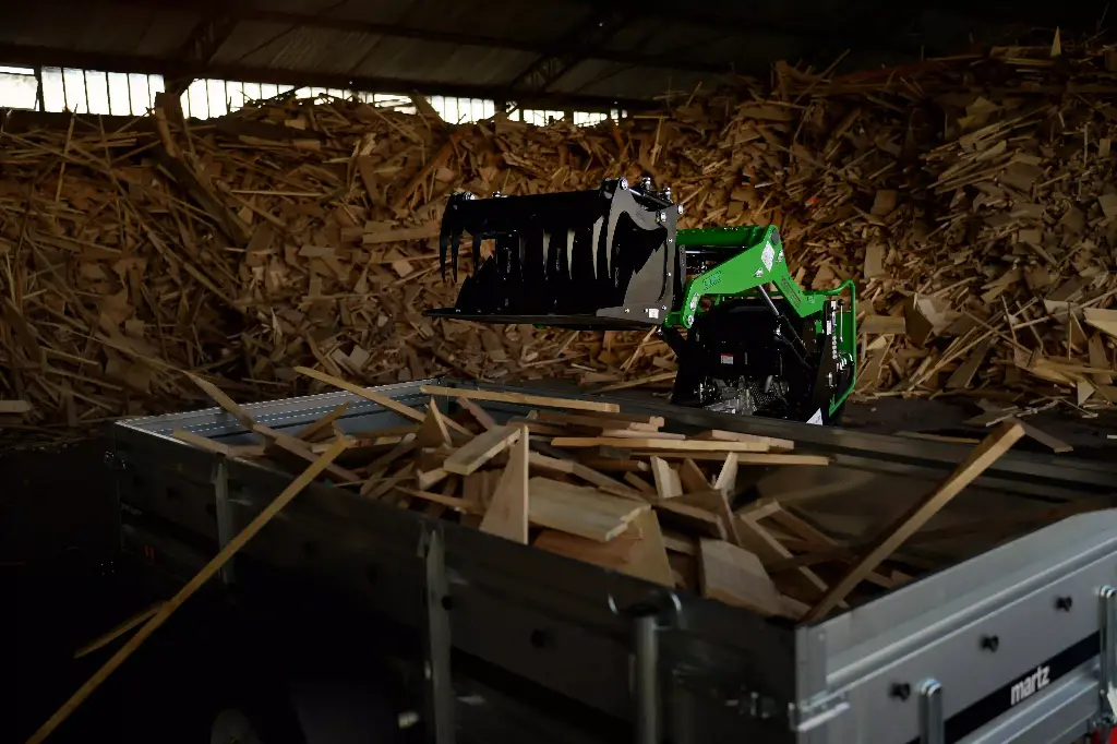 Chargeurplus vous présente le mini chargeur sur chenilles équipé d'un grappin à dents en action dans un hangar, chargeant des chutes de bois au-dessus d'une remorque, devant un imposant tas de déchets de bois, démontrant son efficacité pour le tri et la manutention de déchets encombrants.