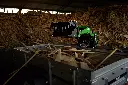 Chargeurplus vous présente le mini chargeur sur chenilles équipé d'un grappin à dents en action dans un hangar, chargeant des chutes de bois au-dessus d'une remorque, devant un imposant tas de déchets de bois, démontrant son efficacité pour le tri et la manutention de déchets encombrants.