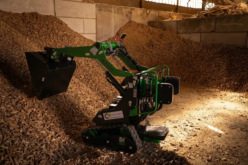 Chargeurplus vous présente le mini chargeur sur chenilles en action dans un bâtiment agricole, bras levé avec godet chargé de copeaux de bois, positionné devant un important tas de broyat, illustrant parfaitement ses capacités de manutention en espace confiné.