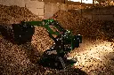 Chargeurplus vous présente le mini chargeur sur chenilles en action dans un bâtiment agricole, bras levé avec godet chargé de copeaux de bois, positionné devant un important tas de broyat, illustrant parfaitement ses capacités de manutention en espace confiné.