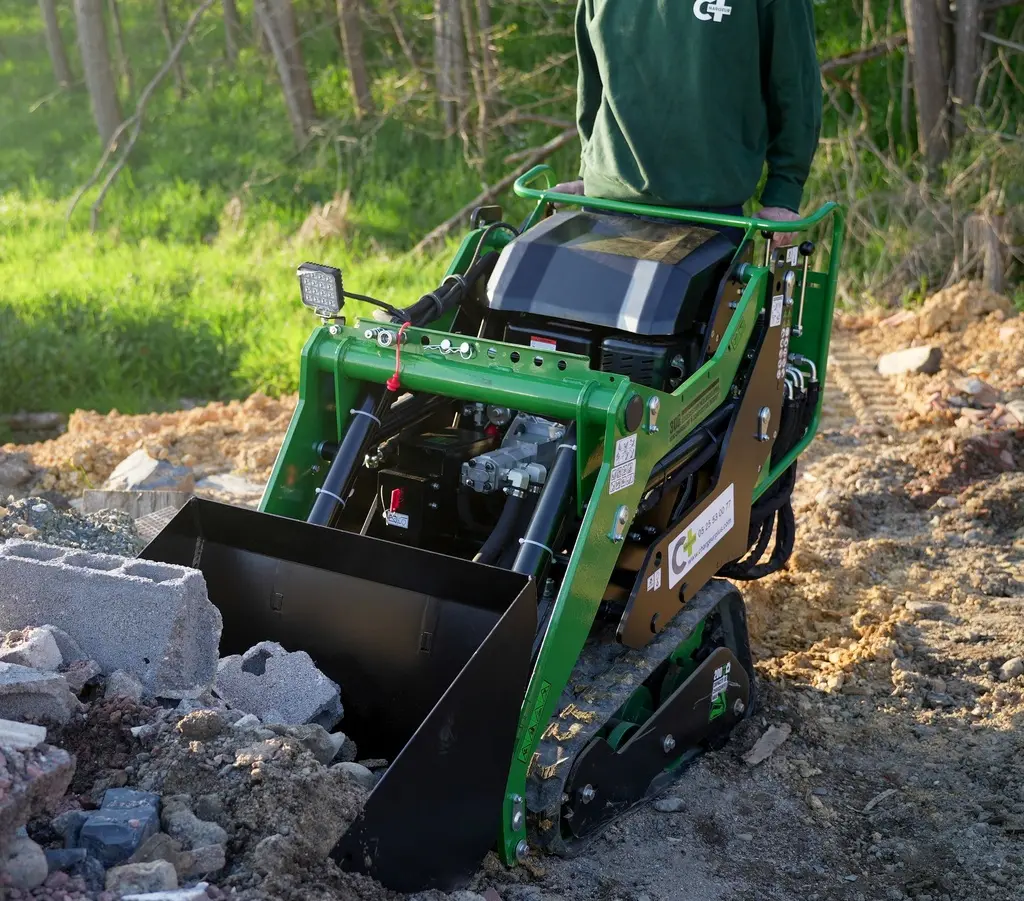 Chargeurplus vous présente un mini chargeur sur chenilles vert en action sur un chantier, conduit debout par un opérateur portant un t-shirt Chargeurplus, godet chargeur abaissé au sol parmi des gravats et parpaings, sur terrain meuble en extérieur.