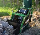 Chargeurplus vous présente un mini chargeur sur chenilles vert en action sur un chantier, conduit debout par un opérateur portant un t-shirt Chargeurplus, godet chargeur abaissé au sol parmi des gravats et parpaings, sur terrain meuble en extérieur.