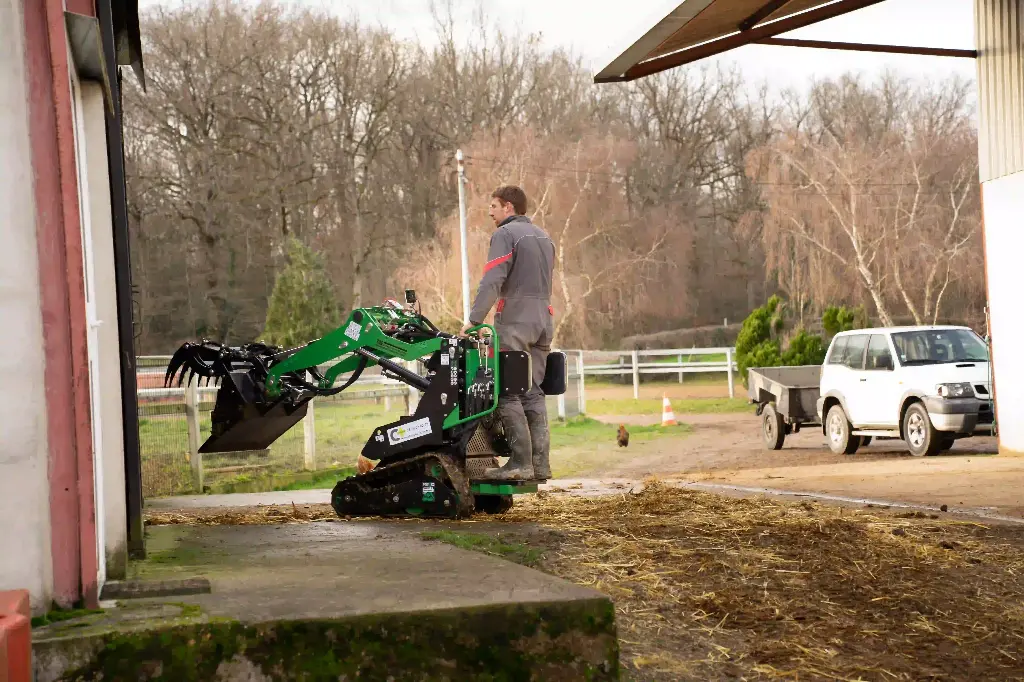 Chargeurplus vous présente le mini chargeur sur chenilles utilisé en milieu agricole, conduit debout par un opérateur à l'entrée d'un bâtiment, équipé d'un godet grappin levé, sur fond de cour de ferme avec véhicule et remorque en arrière-plan.