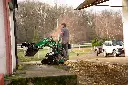 Chargeurplus vous présente le mini chargeur sur chenilles utilisé en milieu agricole, conduit debout par un opérateur à l'entrée d'un bâtiment, équipé d'un godet grappin levé, sur fond de cour de ferme avec véhicule et remorque en arrière-plan.