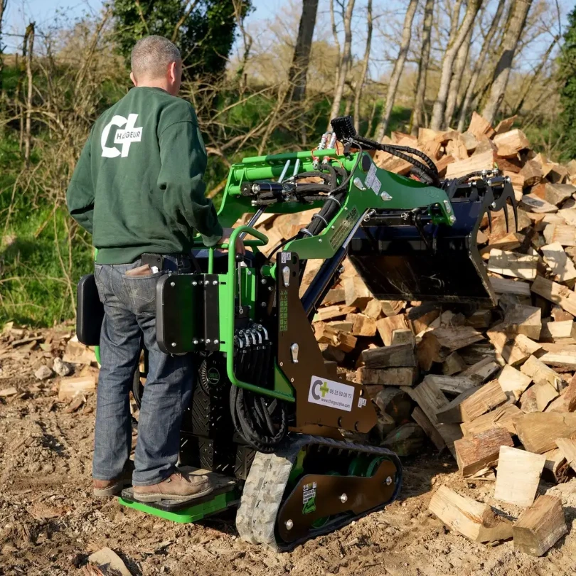 Chargeurplus vous présente le mini chargeur sur chenilles équipé d'un grappin en train de manipuler des bûches de bois de chauffage, conduit debout par un opérateur en sweat Chargeurplus, illustrant parfaitement son efficacité pour la gestion de bois en extérieur.