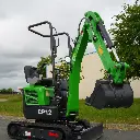 photo de côté d'une mini-pelle CP12 sur une route, on aperçoit sur la photo les chenilles de la mini-pelle, les manettes, et un godet au bout du bras de la flèche. Le vérin est inversé