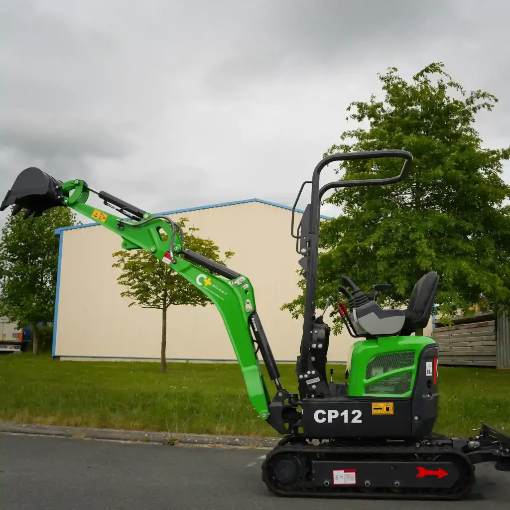 photo de côté d'une mini-pelle CP12 sur une route, on aperçoit sur la photo les chenilles de la mini-pelle, les manettes, et un godet au bout du bras de la flèche. Le vérin est inversé. Le bras est étendu à sa hauteur maximale