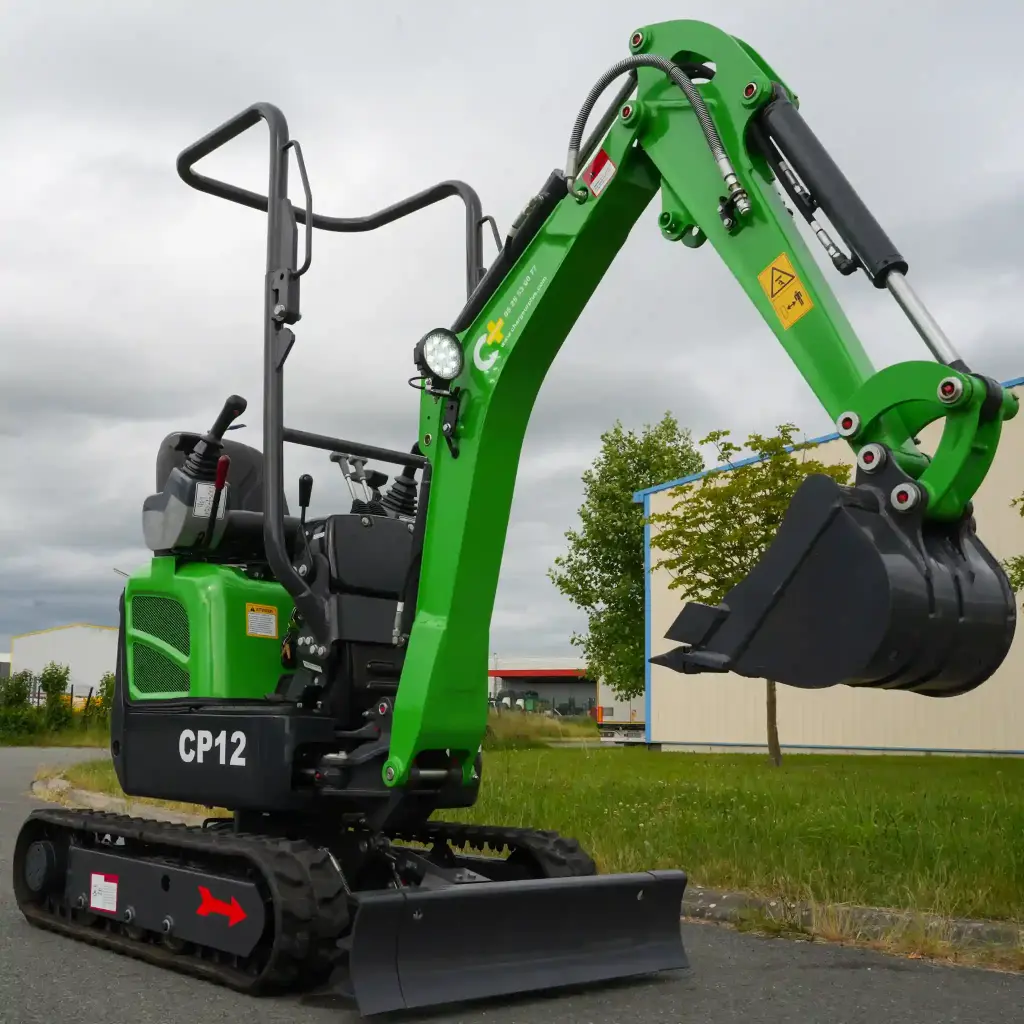 photo de côté d'une mini-pelle CP12 sur une route, on aperçoit sur la photo les chenilles de la mini-pelle, les manettes, et un godet au bout du bras de la flèche. Le vérin est inversé